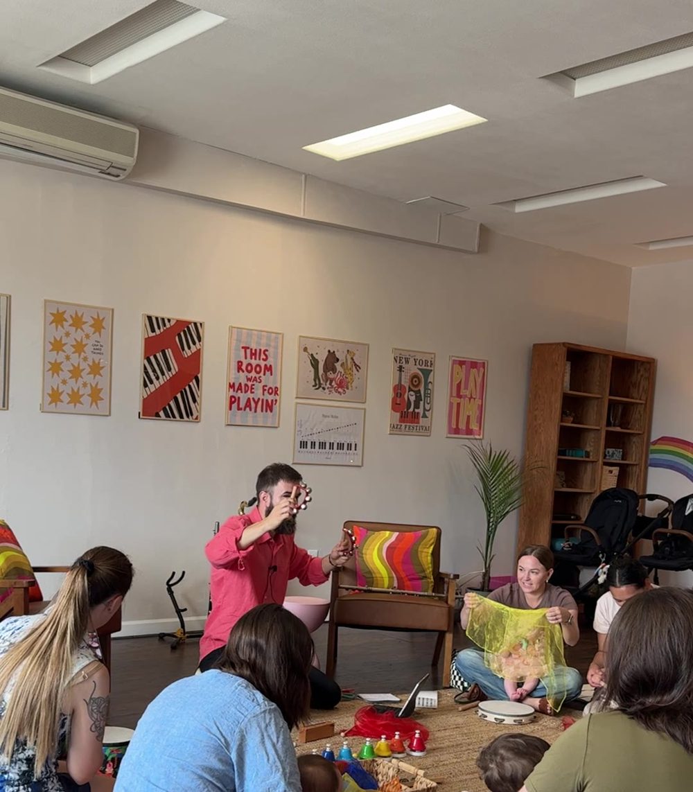 A full circle of families in a Journeys class, with Claudiu leading in front of the 'This room was made for playin'' poster
